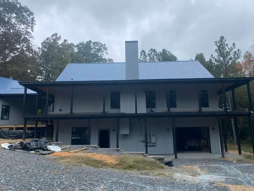 Two-story gray house with black trim, blue metal roof, and covered porch under construction, set in a wooded area.
