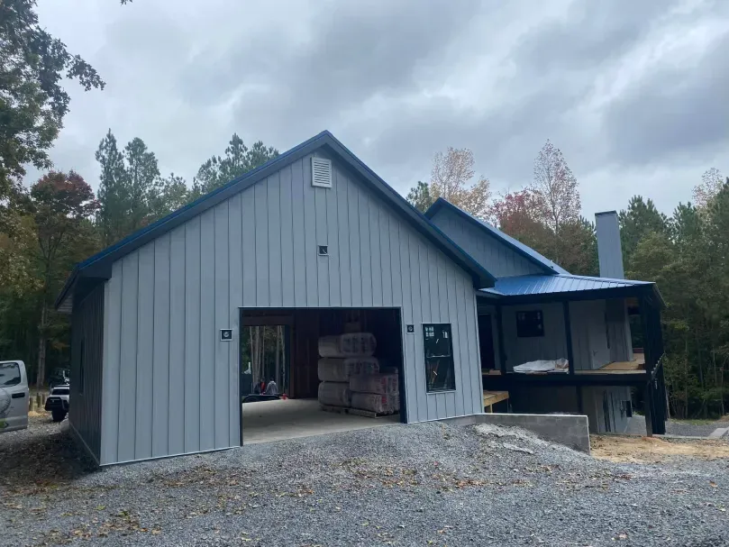 Gray barn-style building with blue roof under construction. Garage and porch visible. Cloudy sky.