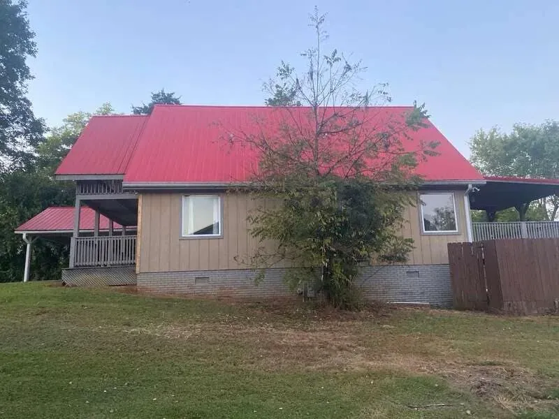 Tan house with red metal roof, a porch, and a brown fence, surrounded by green grass and trees.
