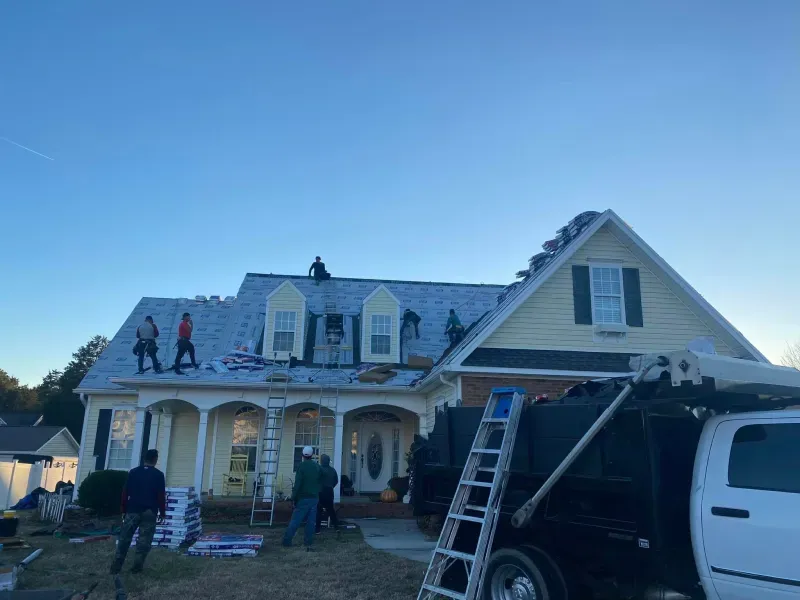 Roofers working on a house, with materials and truck nearby. Blue sky, daylight.