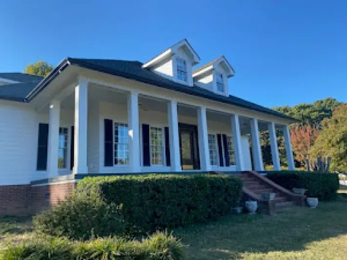White house with columns, black shutters, and dormer windows, on a sunny day.