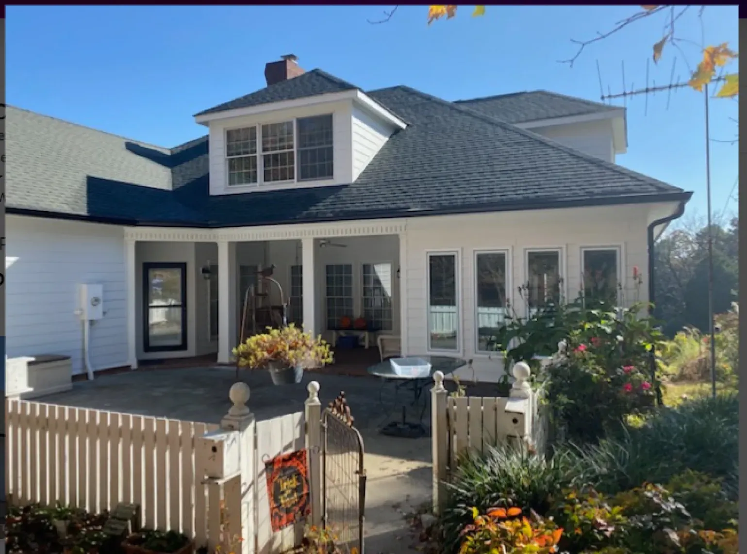 White house with dark roof and porch, surrounded by a picket fence and greenery on a sunny day.