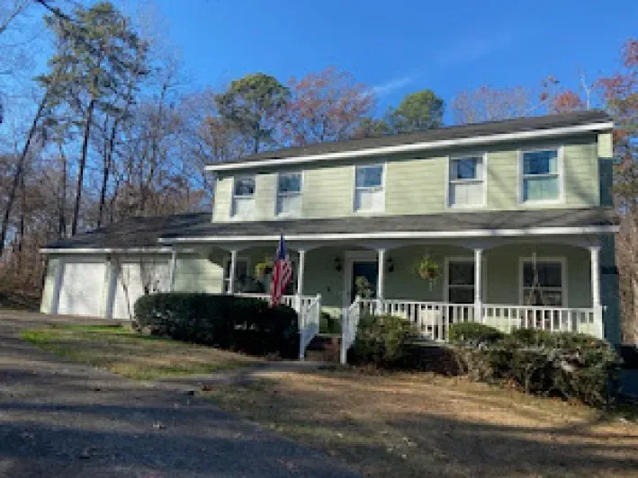 Two-story light green house with white porch, two-car garage, and American flag on a sunny day.