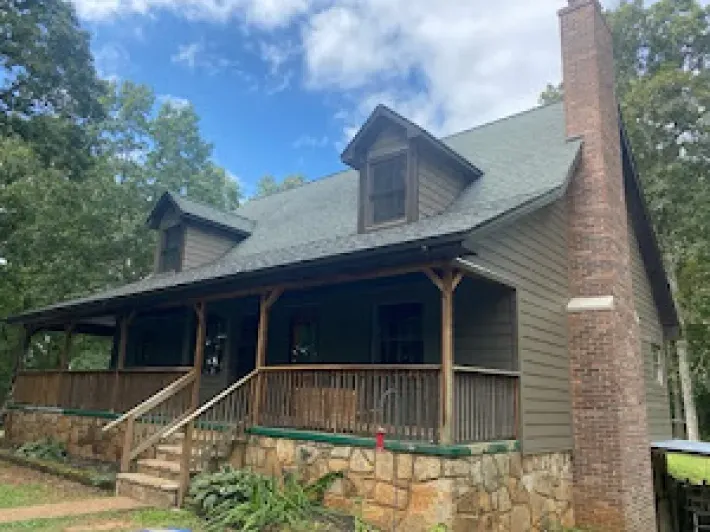 A two-story house with a wraparound porch and a stone foundation, set against a background of trees.