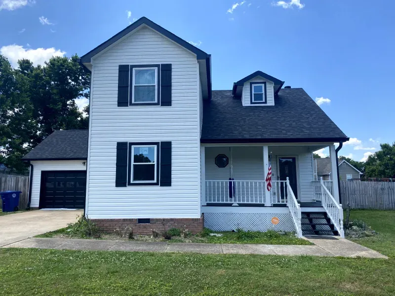 White two-story house with black trim, shutters, and garage door. A porch and dormer are visible; green lawn.