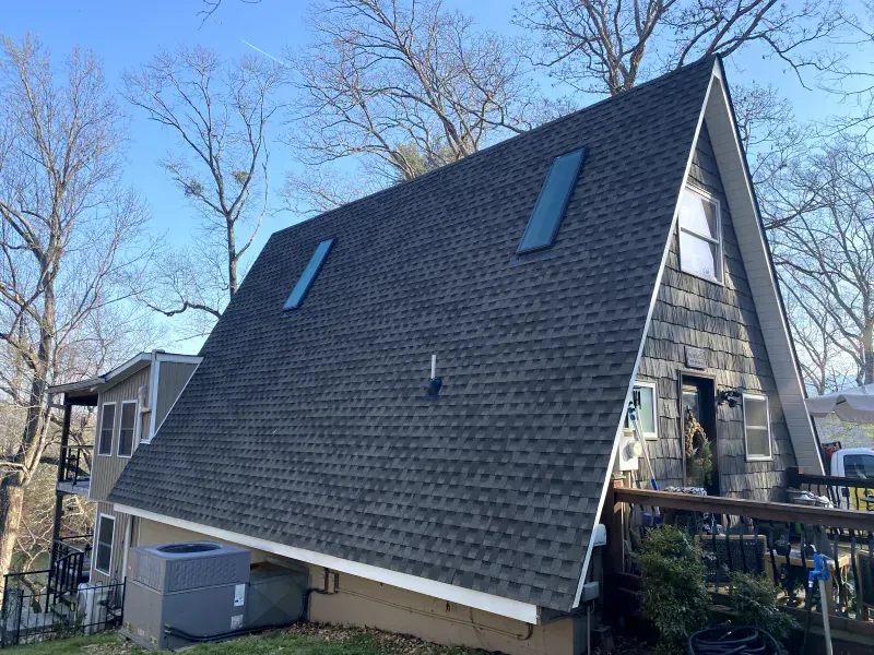 A-frame house with a dark gray shingle roof, small windows, and a deck, set among bare trees on a sunny day.