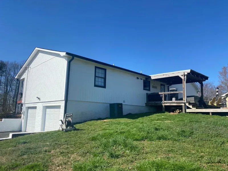 White house with black trim, two-car garage, and deck on a grassy hill under a blue sky.