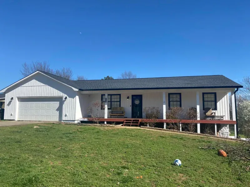 White house with a dark roof and porch, set against a bright blue sky and green lawn.