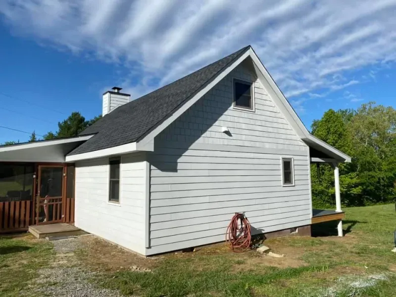 Gray-sided house with a dark roof and a porch. A hose and chimney are visible, set on a grassy area under a bright sky.
