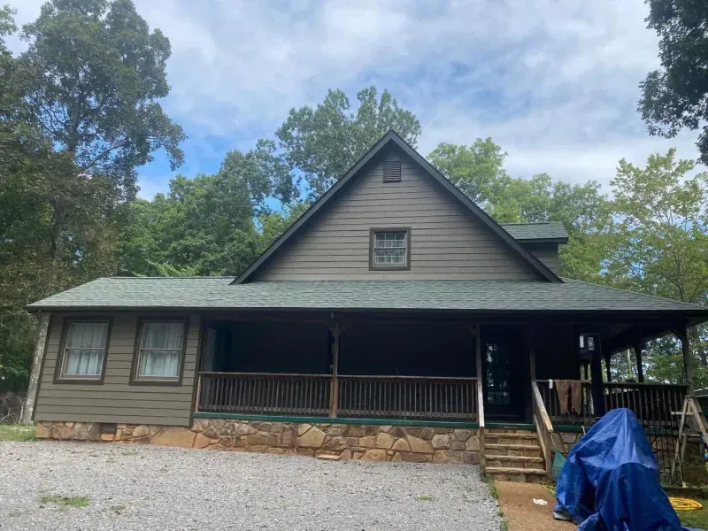 A one-story brown house with a porch and stone foundation, surrounded by trees under a cloudy sky.