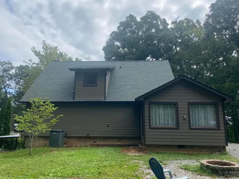 Brown cabin with a dark green roof, surrounded by trees and grass, under a cloudy sky.
