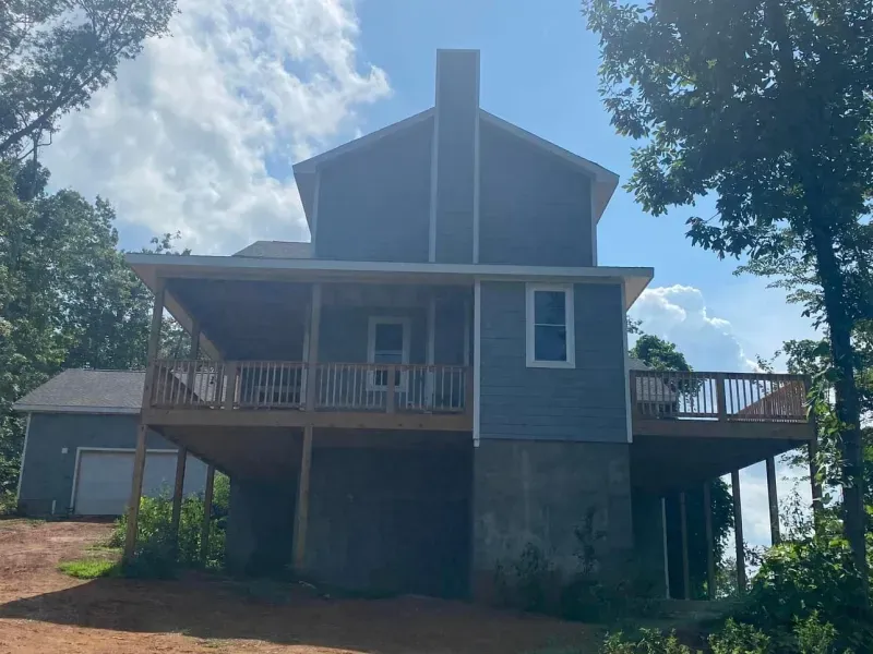 Blue house with wooden deck, attached garage, and chimney, set against a cloudy sky.