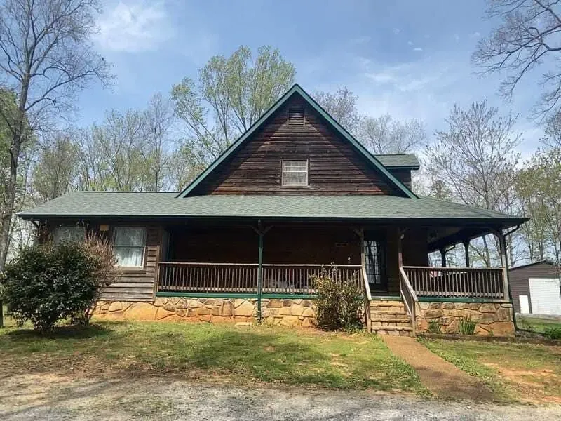 Wooden cabin with porch, brown exterior, green roof. Trees in background, cloudy sky.