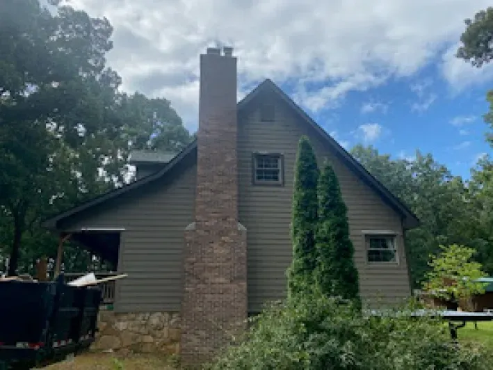 A cabin with a tall brick chimney. Gray siding and a cloudy blue sky. Green trees surround it.