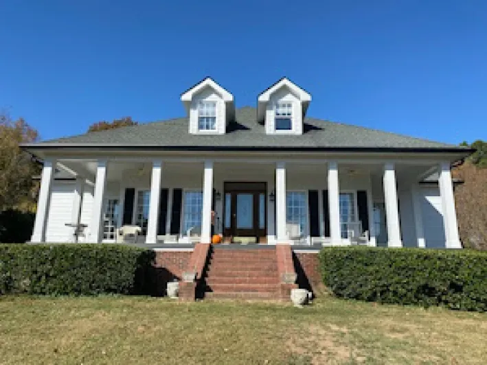 White house with pillars, black shutters, brick steps, and two dormers under a blue sky.