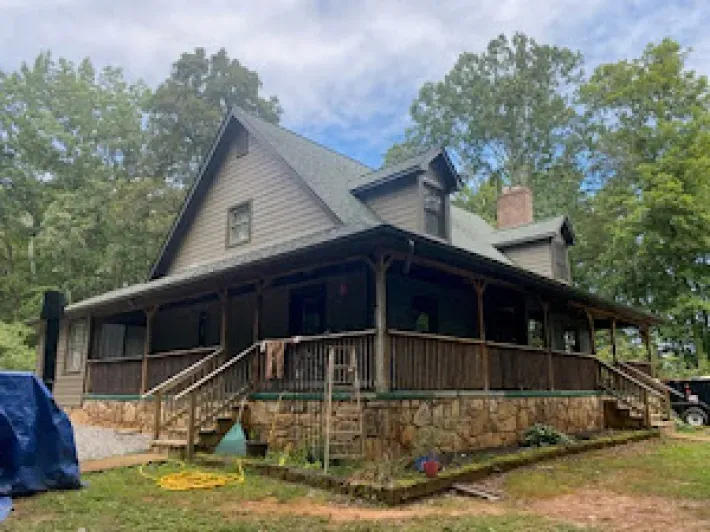 Two-story house with wraparound porch and stone foundation. Green roof, brown siding, and surrounded by trees.