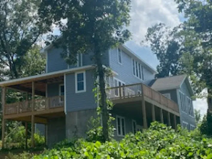Two-story blue house with wooden decks surrounded by green trees.