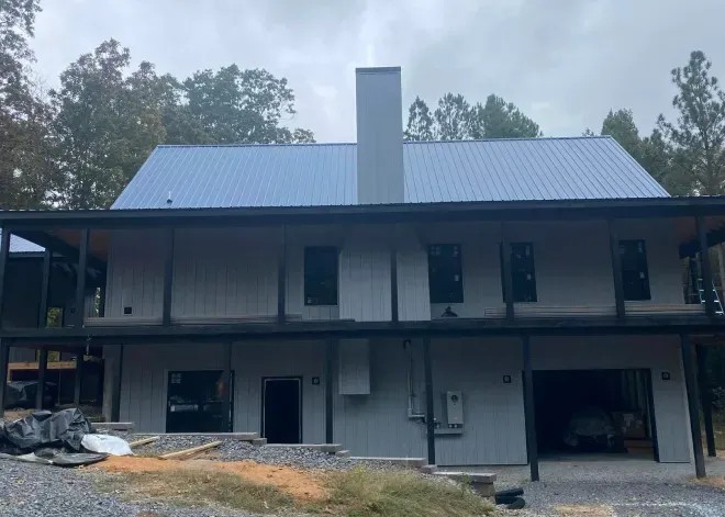 Two-story modern house under construction with a metal roof and garage. Gray exterior, surrounded by trees.