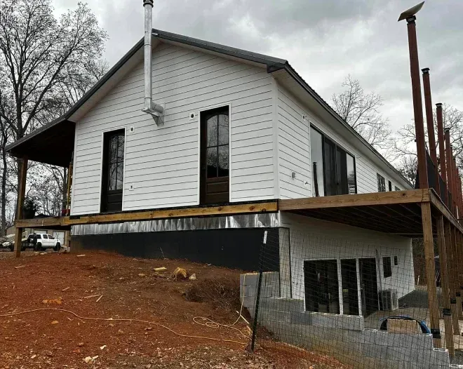 White house with dark windows and exposed wooden deck under construction on a hillside.