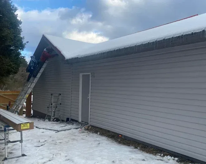 Two people on a ladder, removing snow from a building's roof with gray siding. Snowy ground.