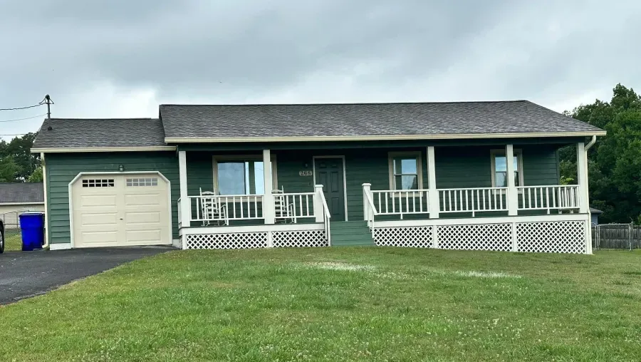 Green house with white porch and garage, on a green lawn. Cloudy sky overhead.
