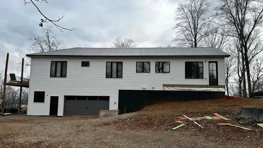 White house with dark windows and garage. Deck on the left, set on a hill with bare trees and cloudy sky.