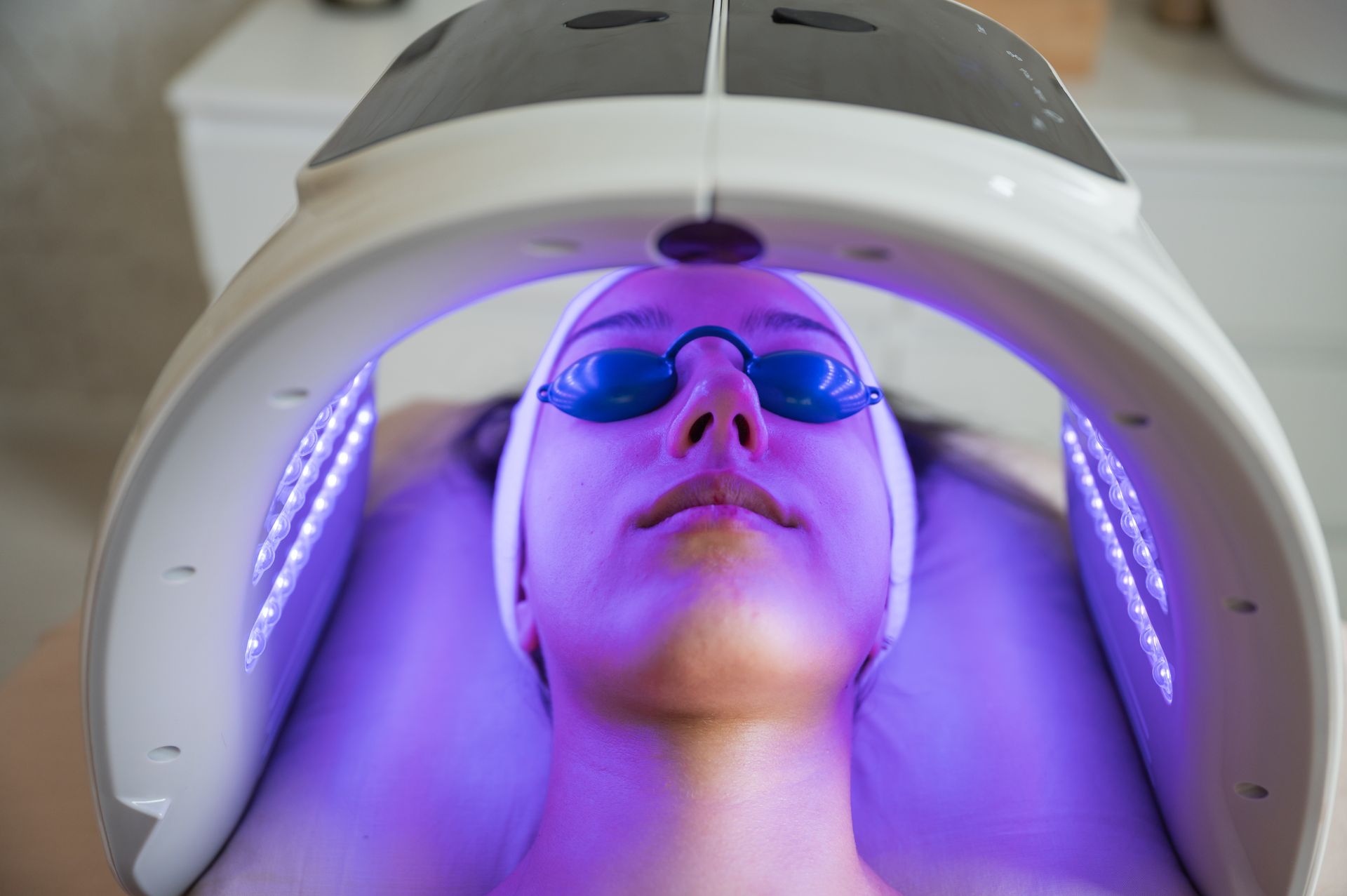 A person lying down wearing blue protective goggles under a curved LED light therapy machine emitting purple light.