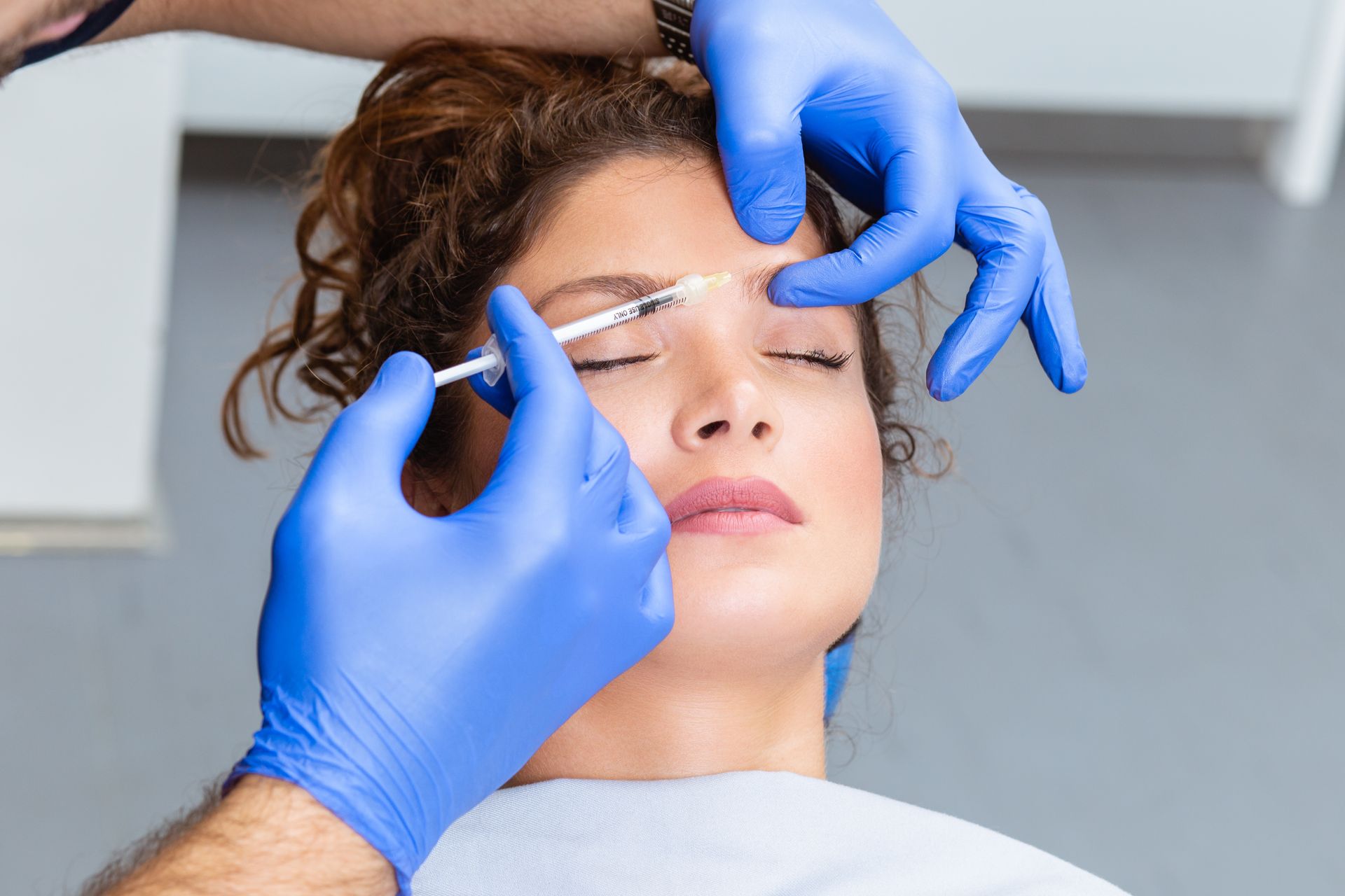 A medical professional in blue gloves administers a facial injection to a person's forehead in a clinical setting.