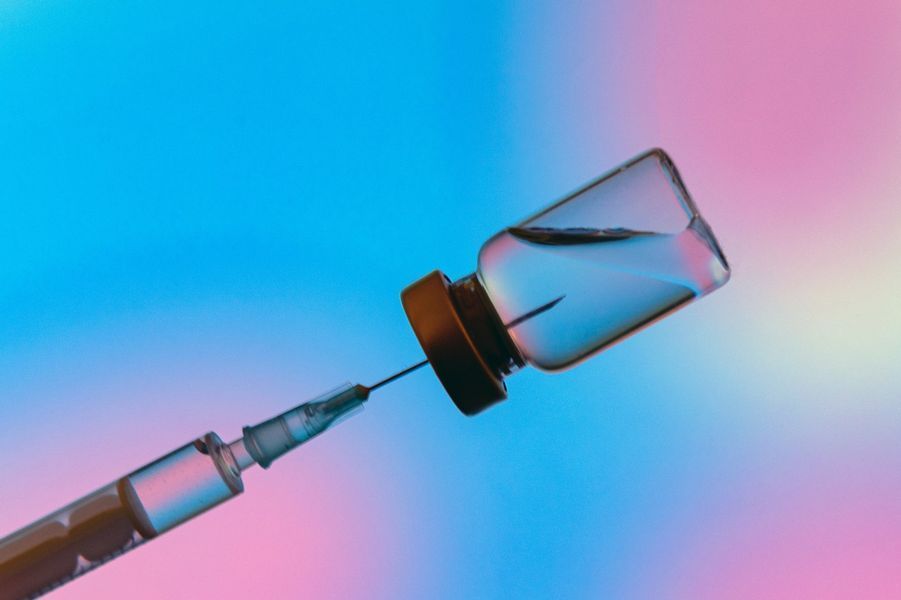 A medical syringe is inserted into a glass vaccine vial against a blurred blue and pink gradient background.