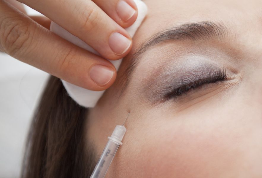 A needle injecting a cosmetic treatment into the side of a person's eye area while a hand holds a cotton pad nearby.