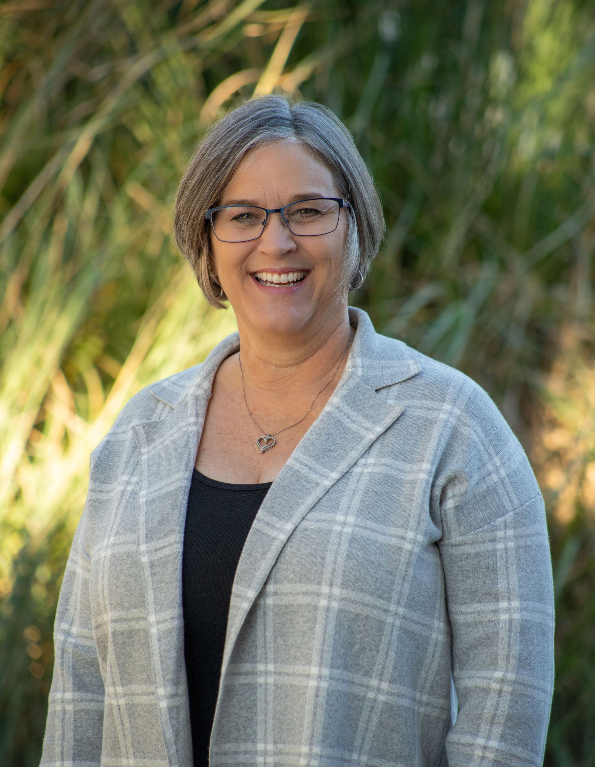 Woman wearing glasses and a plaid jacket smiles, with a background of green foliage.