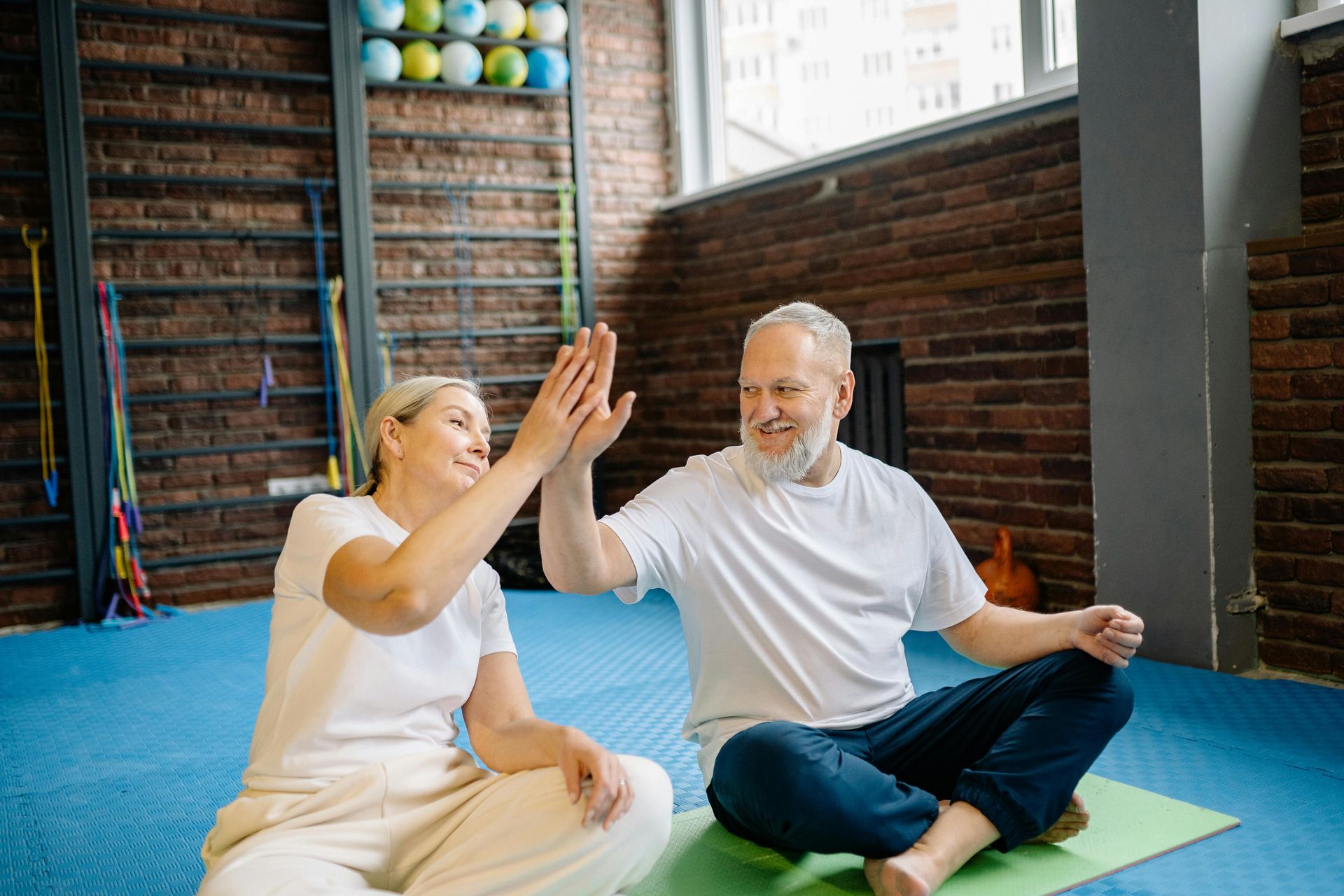 An elderly couple is giving each other a high five while sitting on a yoga mat.