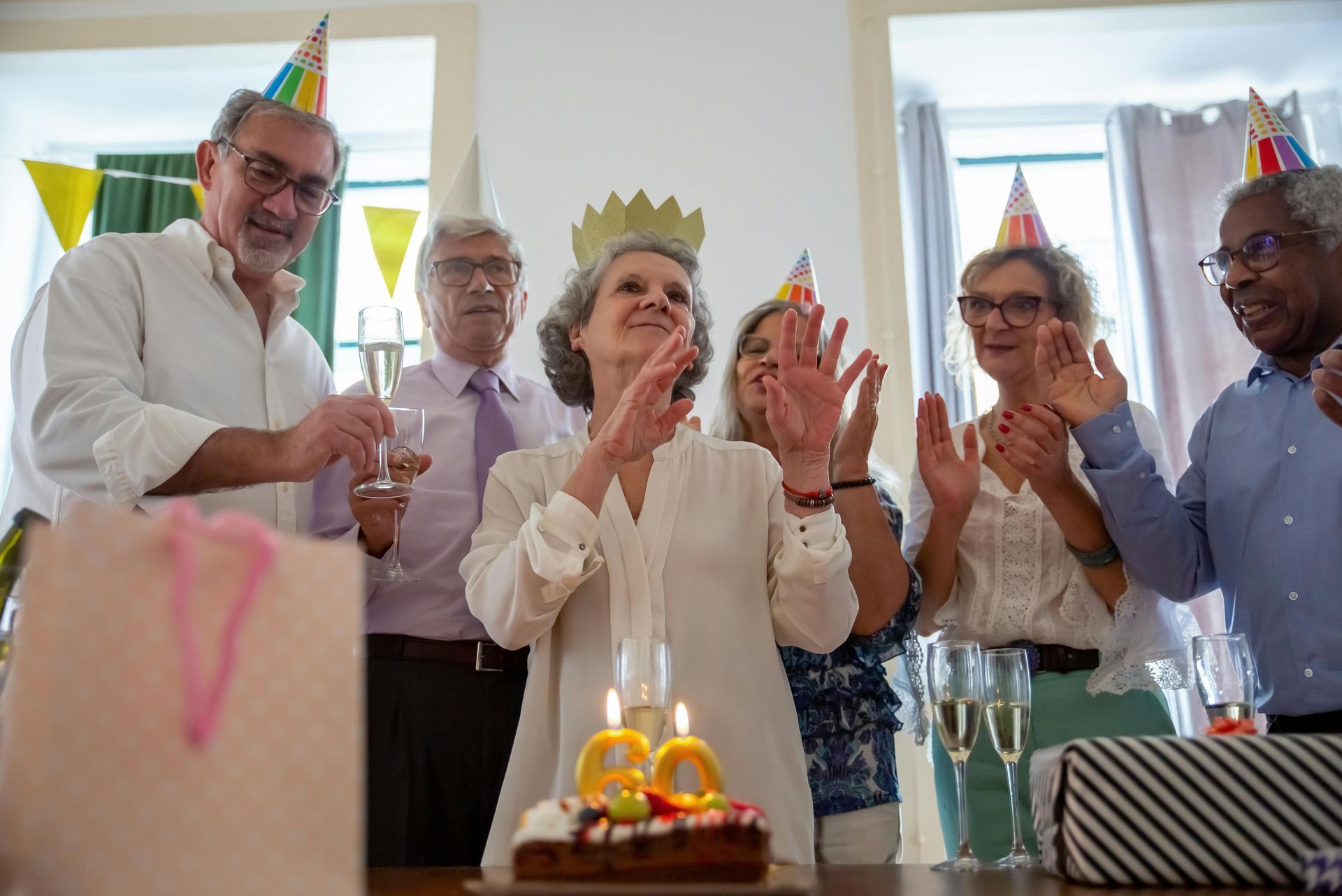 An elderly woman is blowing out candles on a birthday cake while a group of people applaud.