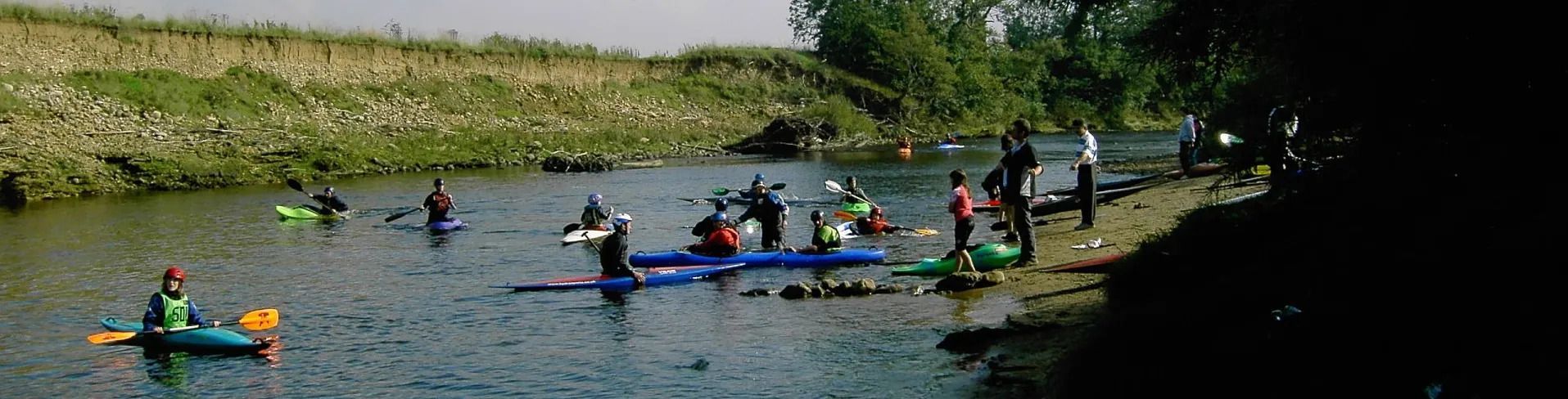 Kayakers on a river, trees on the right, grassy bank on the left, sunny day.