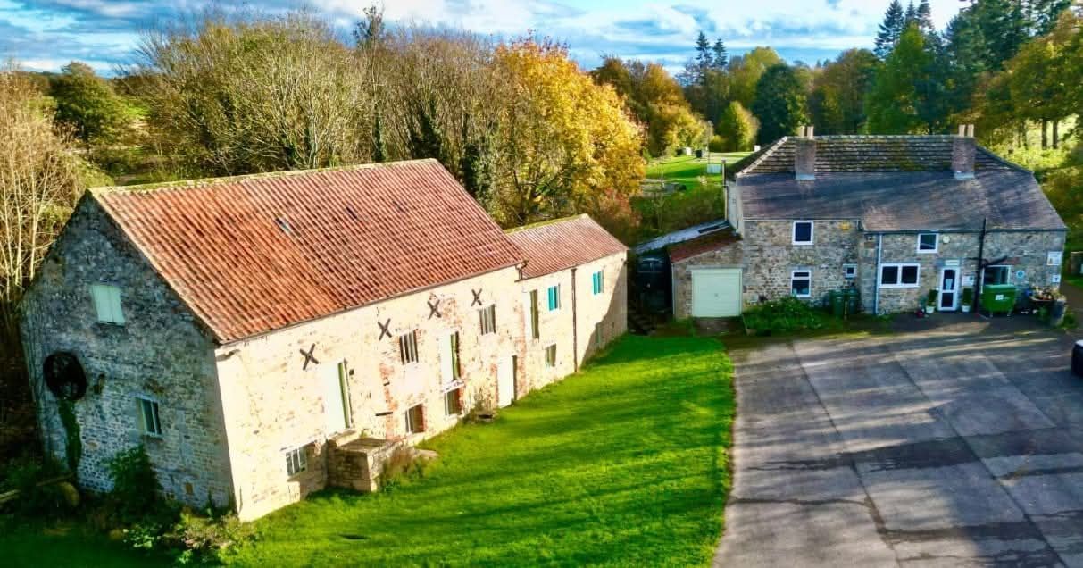 Stone buildings: barn with red roof and farmhouse, green lawn, trees, and parking.