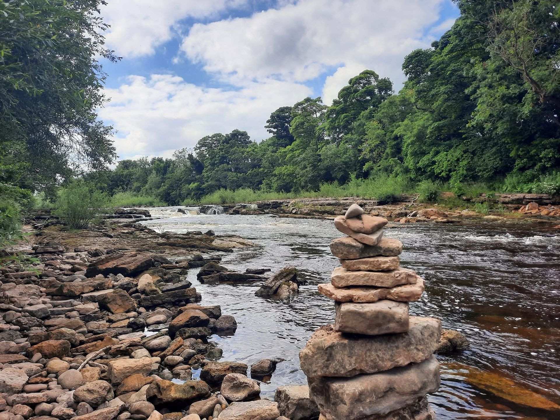 A stack of rocks sits on a rocky riverbank, the river flowing towards a wooded area under a cloudy sky.