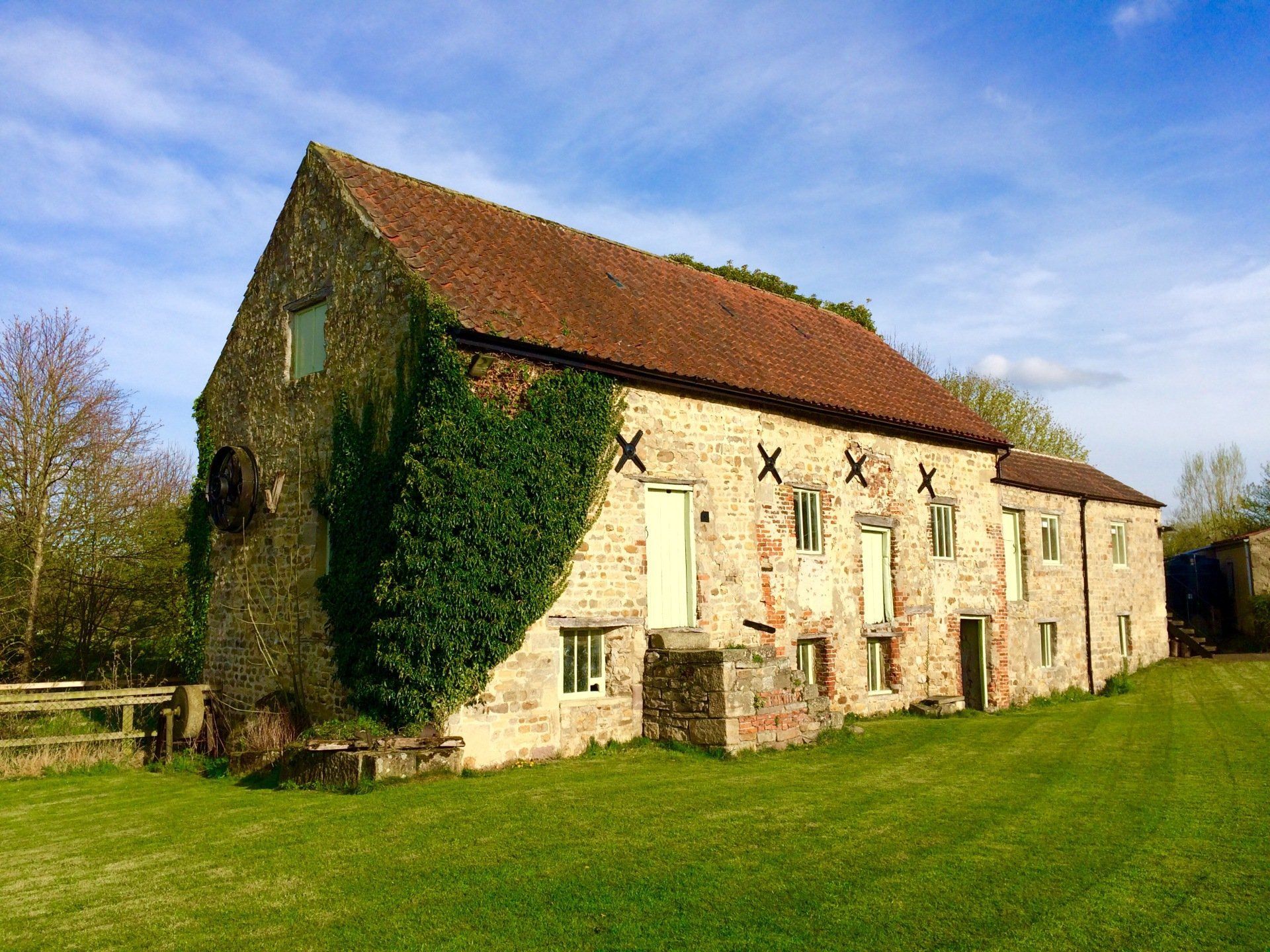 Stone barn with red roof and green ivy, set in a grassy field under a blue sky.