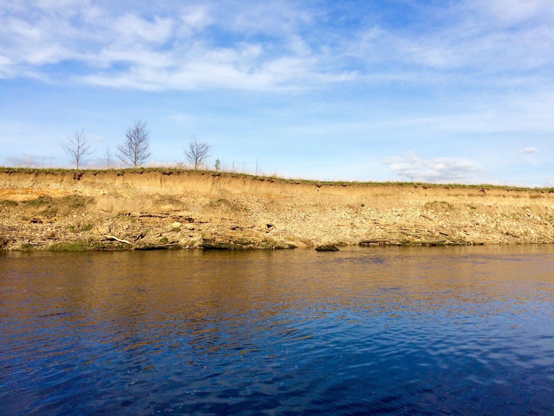 Riverbank with a sandy cliff, calm water, and blue sky with clouds.