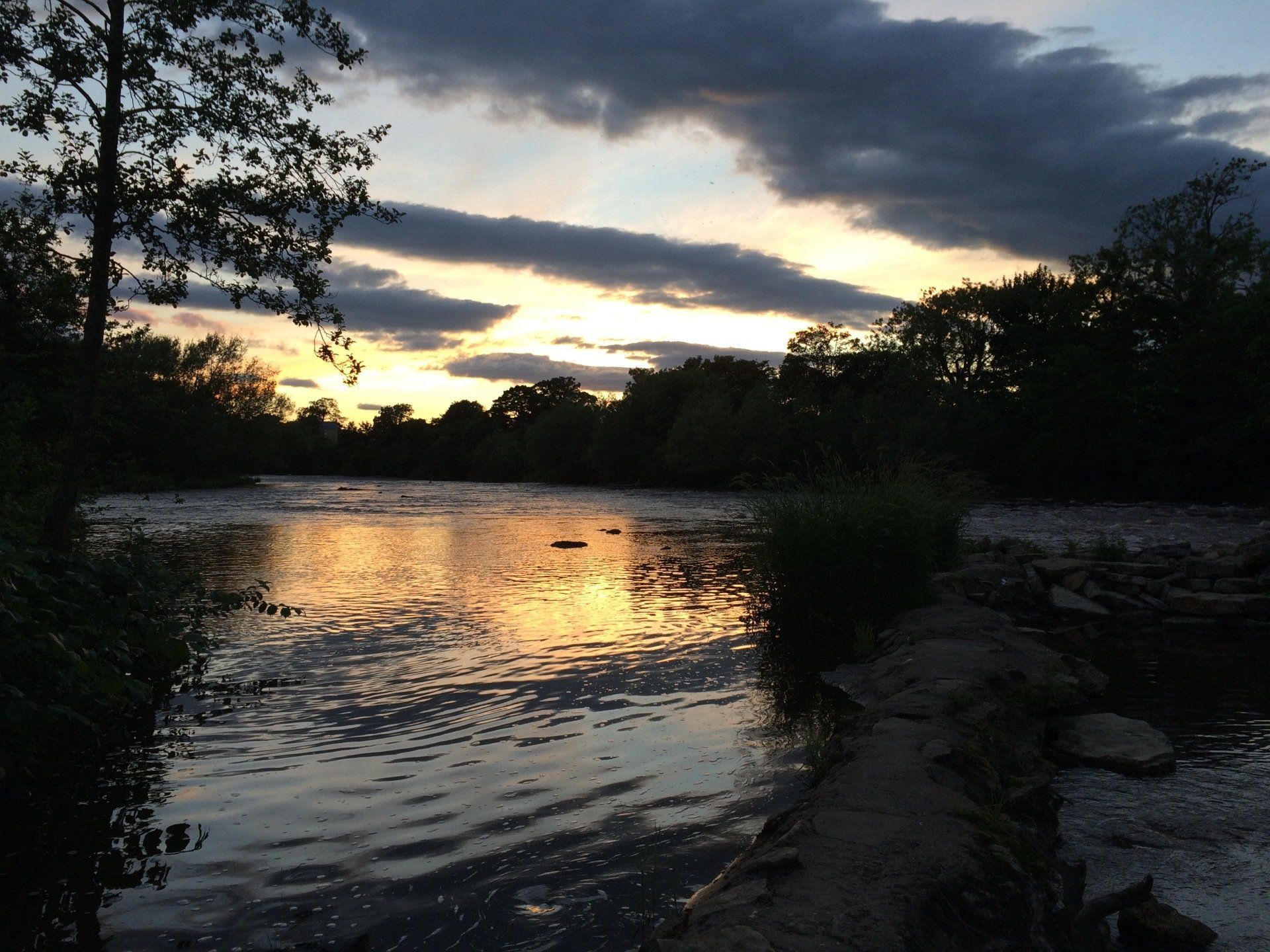 Sunset over a calm river reflecting orange and yellow light; trees line the banks under a cloudy sky.