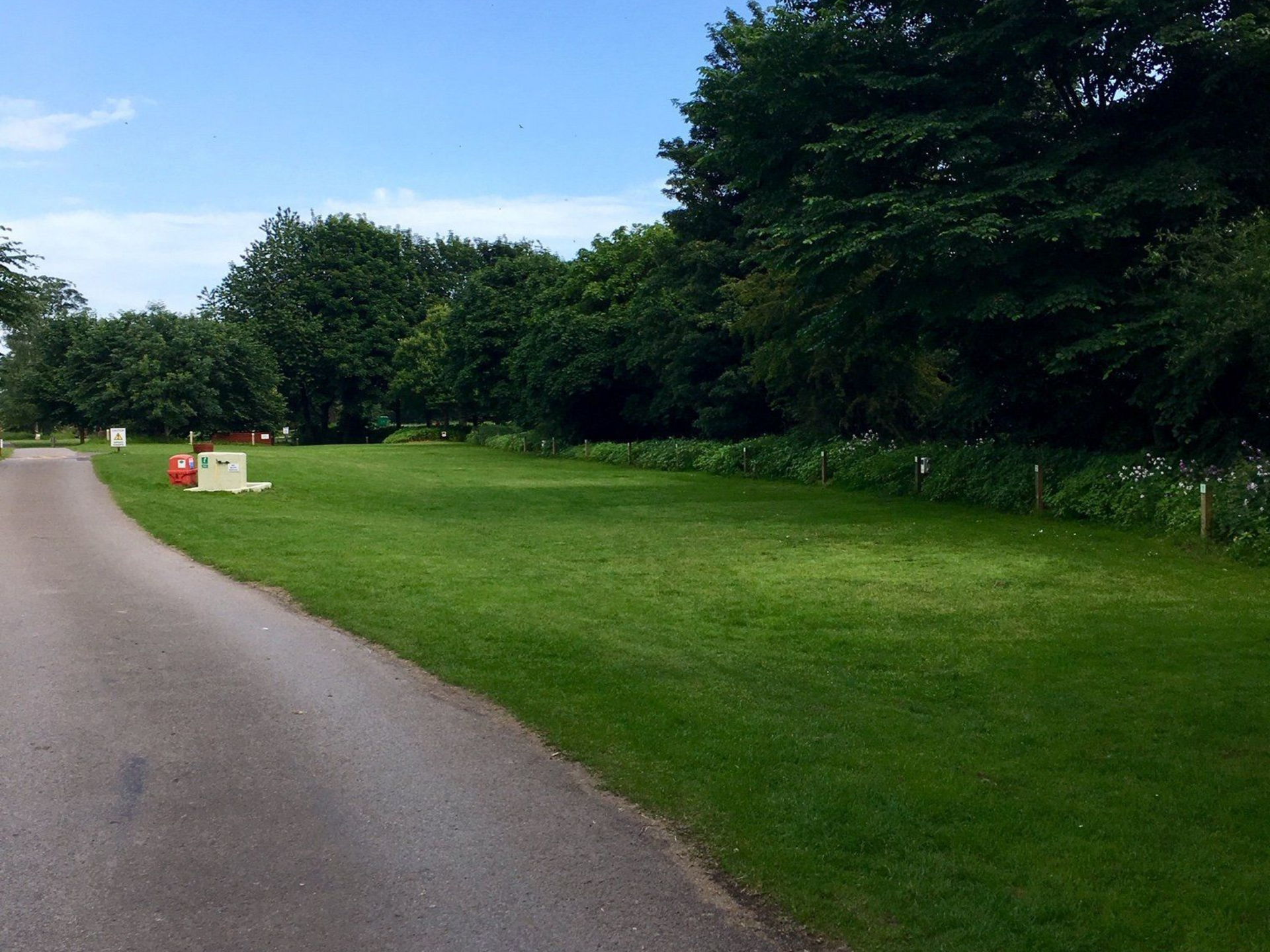 Paved road next to grassy area with trees and a red box on the left, under a blue sky.