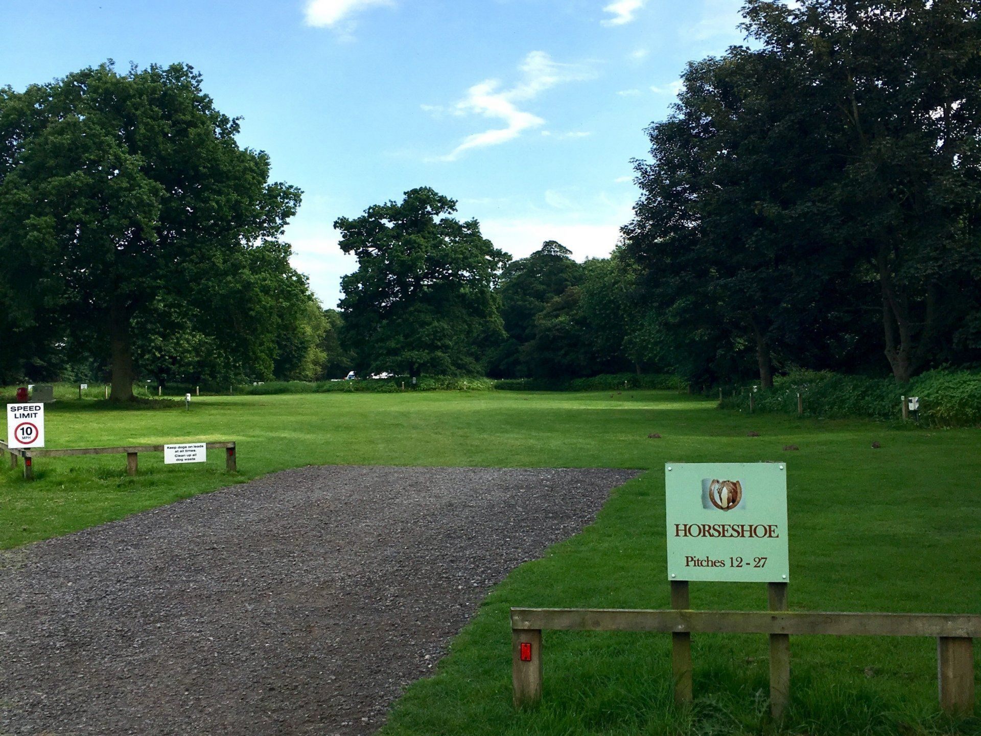A gravel path leads to a grassy park with trees under a partly cloudy sky. Signs are visible.