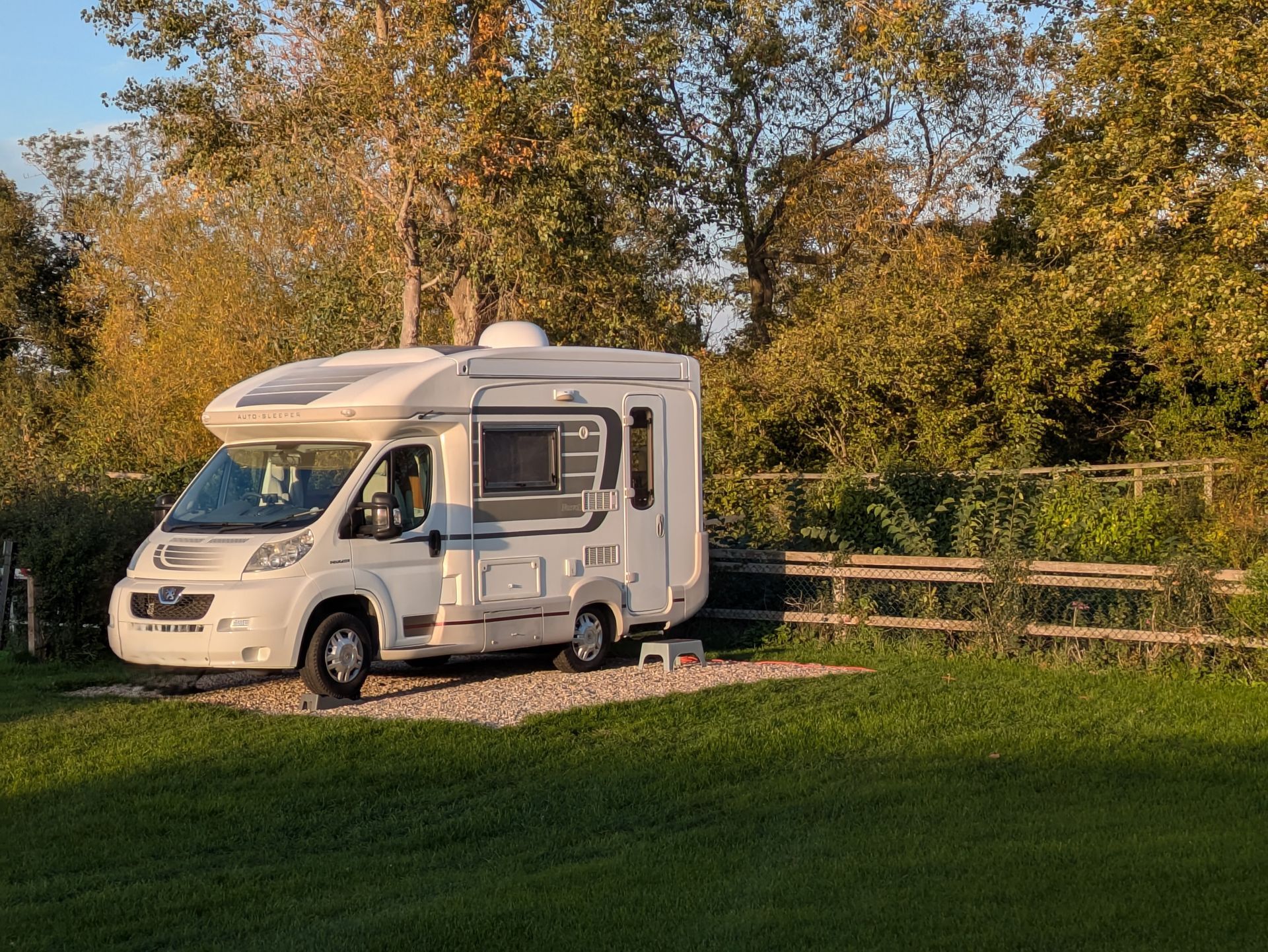 White RV parked on gravel at a campsite, surrounded by grass and trees; golden hour sunlight.