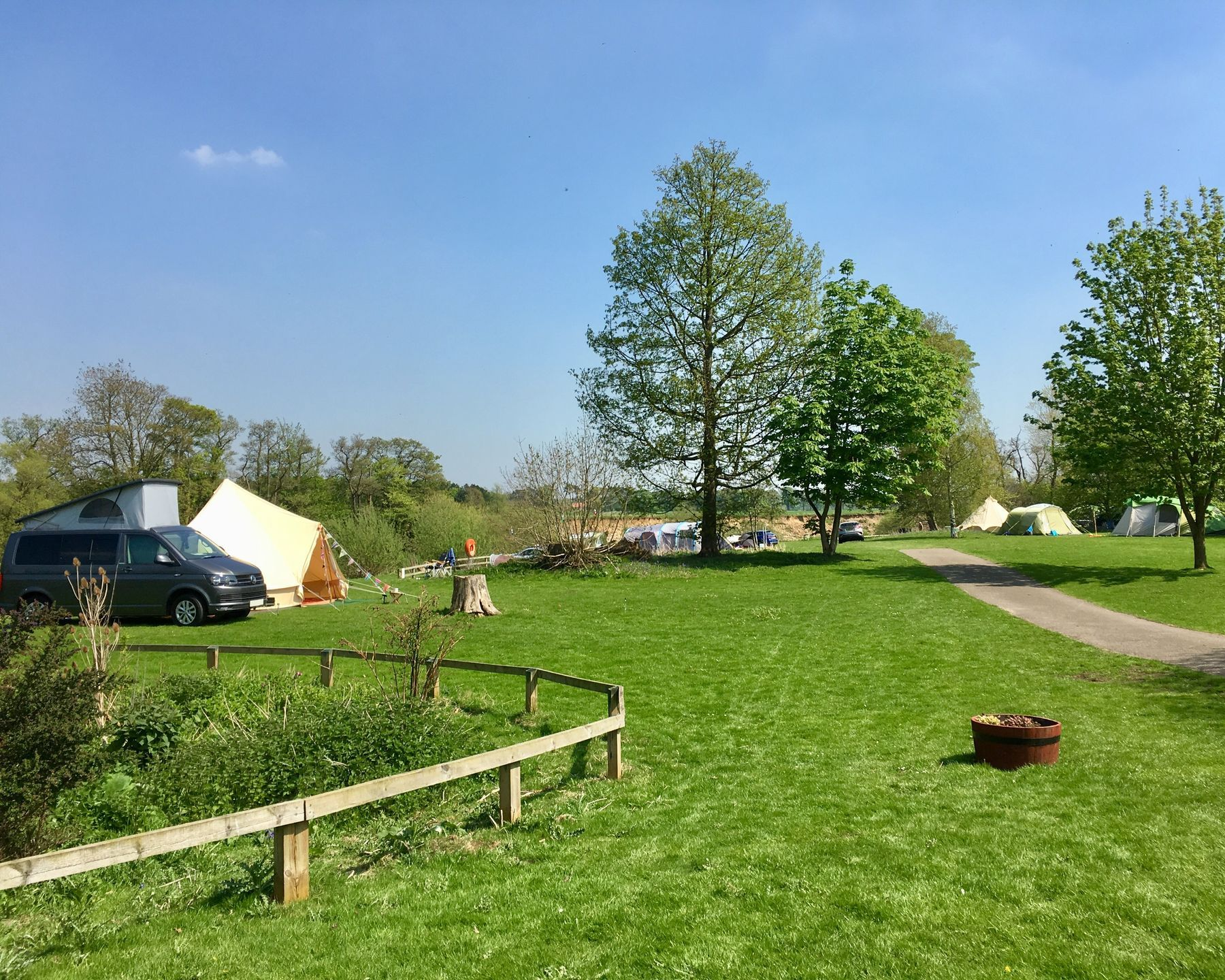 Camping field with tents, van, trees, and a path. Sunny, green grass.