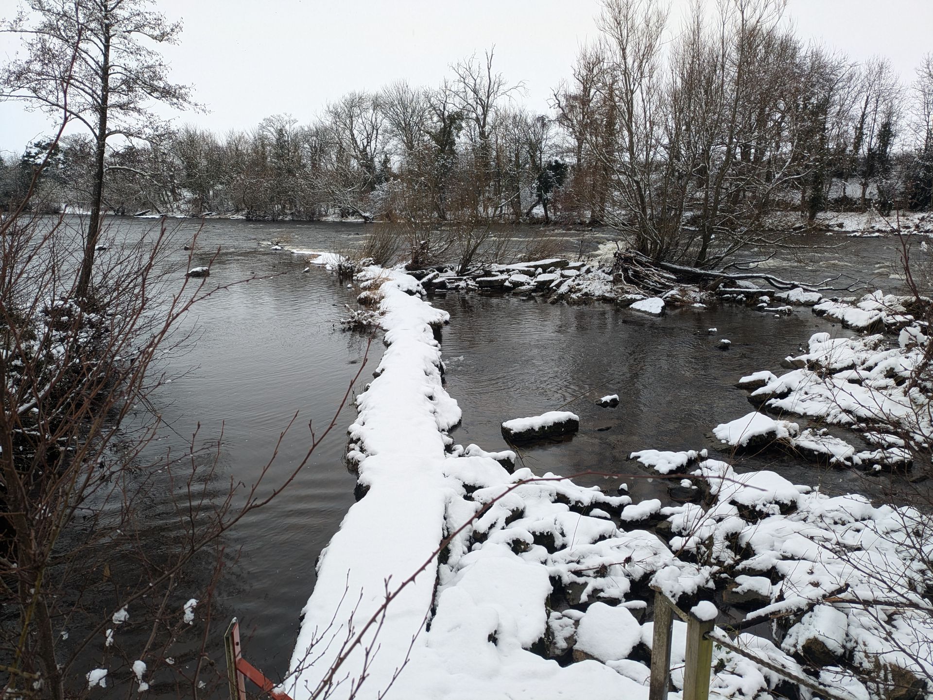 Snow-covered stone causeway across a river, trees and bushes line the banks. Winter scene.