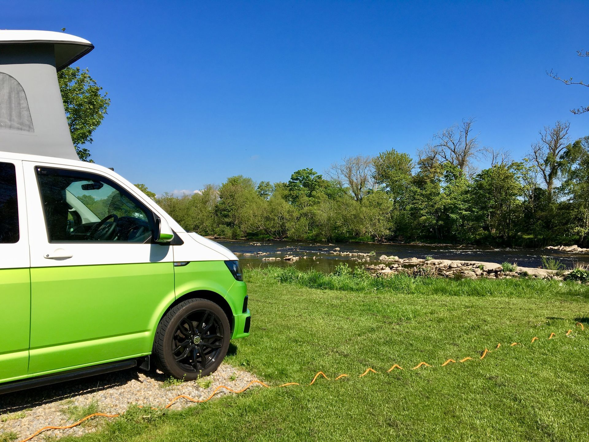 Green and white camper van parked next to a river with a blue sky.