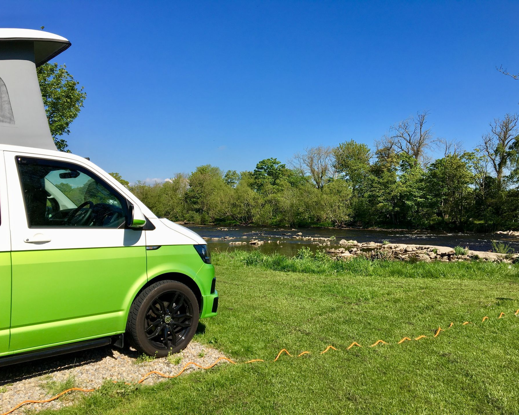Green and white camper van parked near a river on a sunny day.