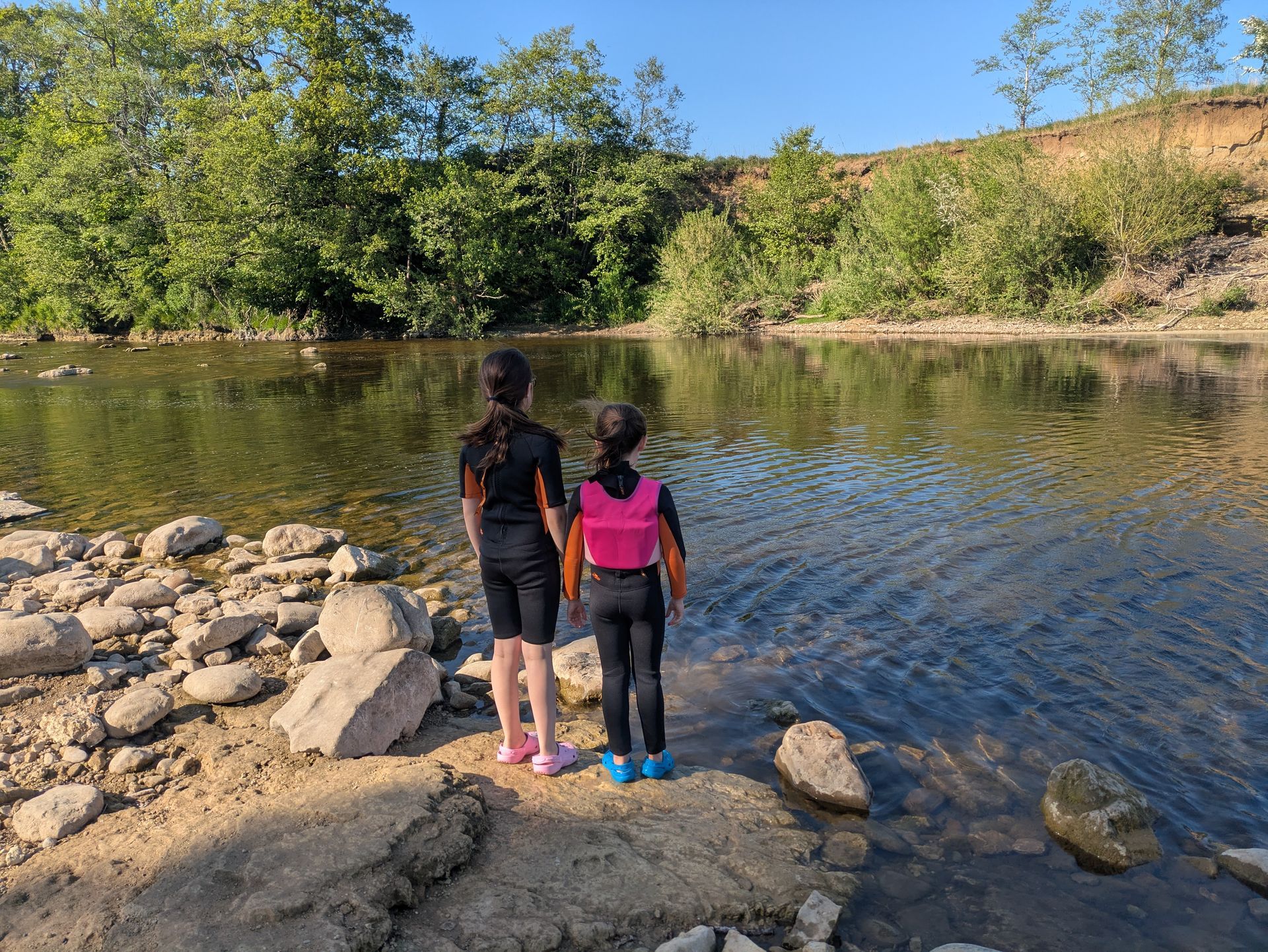 Two people in swimwear stand at the edge of a calm river, looking out over the water.