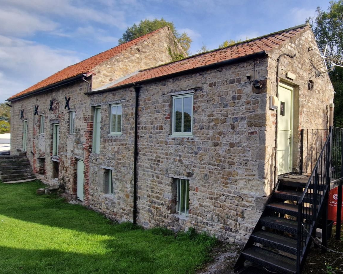 Stone building with red roof, green doors and windows, and a black staircase on a grassy lawn.