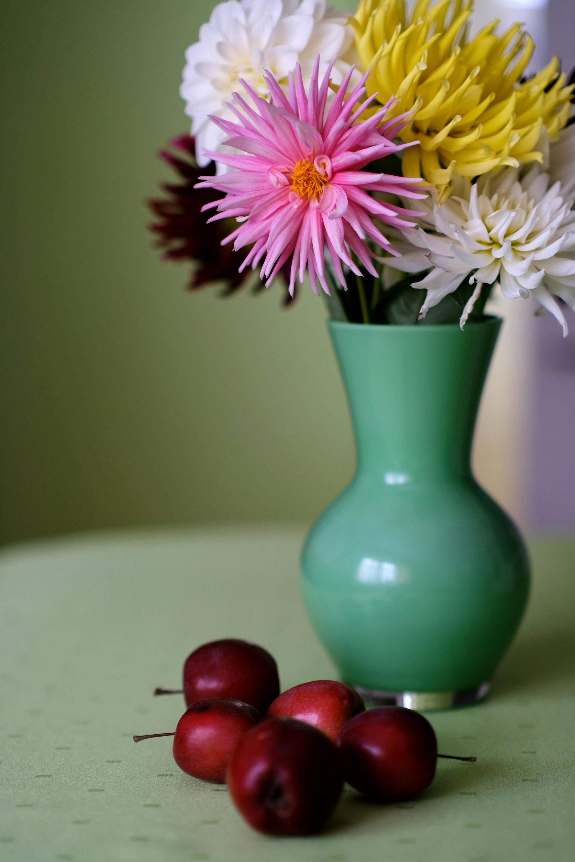 Green vase with colorful dahlias and small red apples on a light green surface.