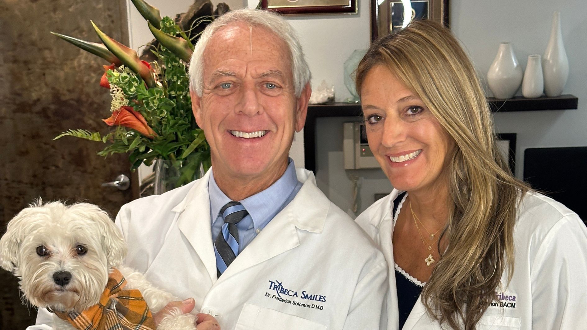 Dr. Frederick Solomon, a cosmetic dentist, and Dr. Danielle Solomon, a licensed acupuncturist, smiling together in white lab coats. Dr. Frederick is holding Oakley, a small white fluffy dog wearing a plaid bow tie, in their Tribeca Smiles office.